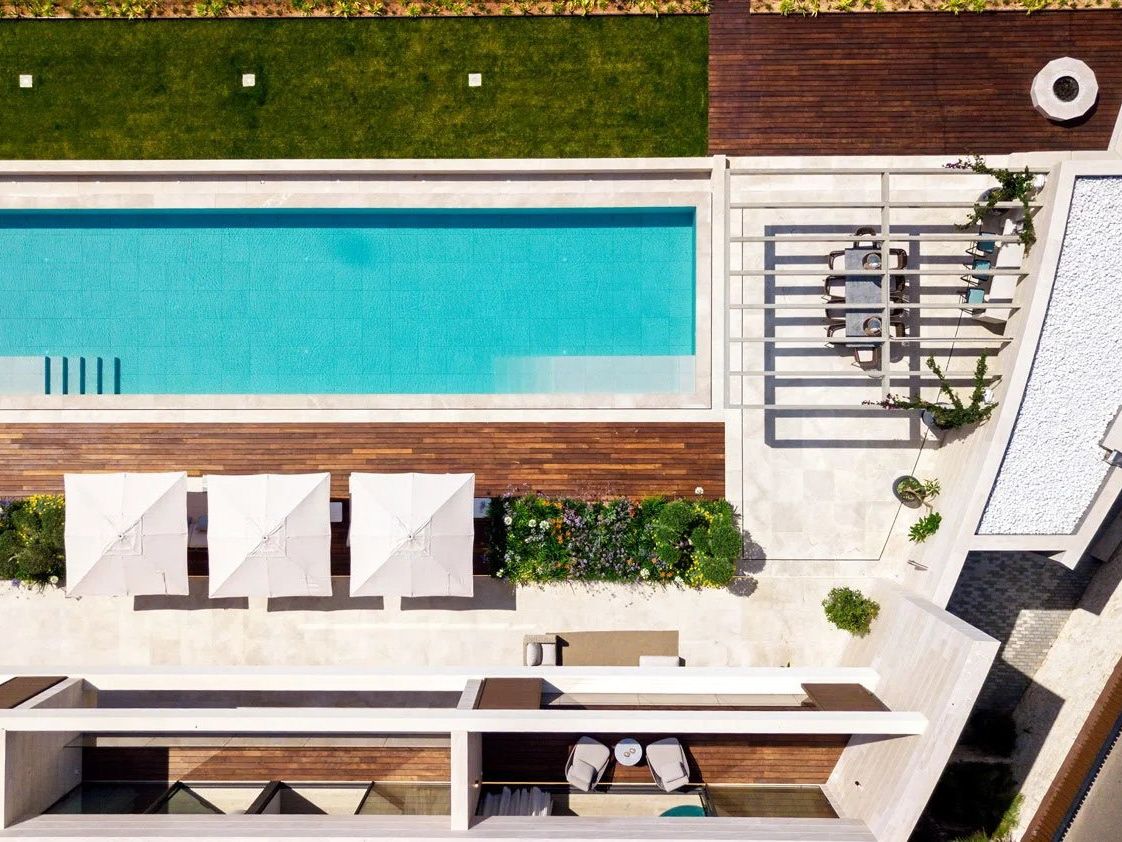 Overhead view of a modern villa terrace — lap pool, timber deck, parasols and a dining setting.