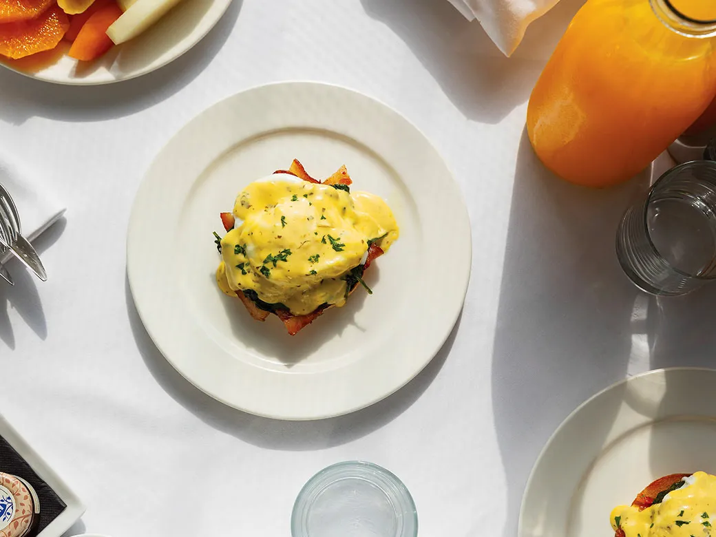 Overhead of eggs benedict on a white plate in hard morning light, fresh juice alongside.