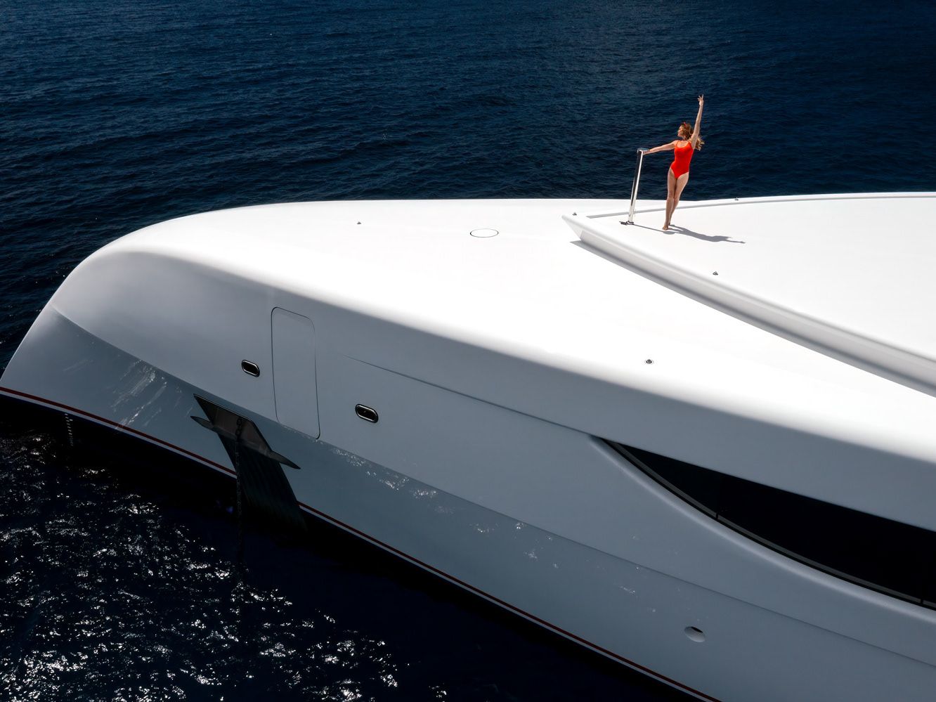 Aerial of a white superyacht at anchor on deep blue open water, a guest standing at the bow.
